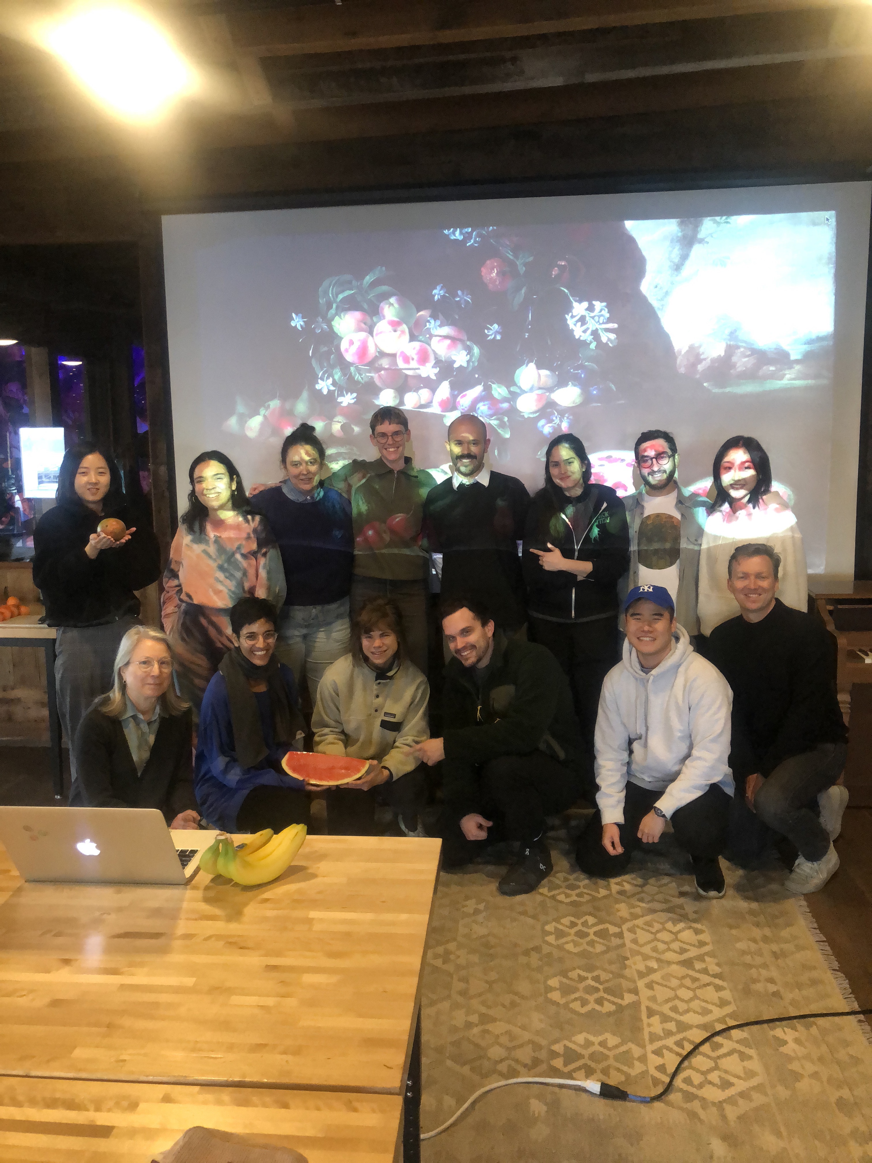 A group of mostly smiling people pose with various fruits and a projection of a renaissance painting of fruit. In the foreground, there's an Apple laptop next to a few bananas on a wooden table and a cable charging trails off to the right.