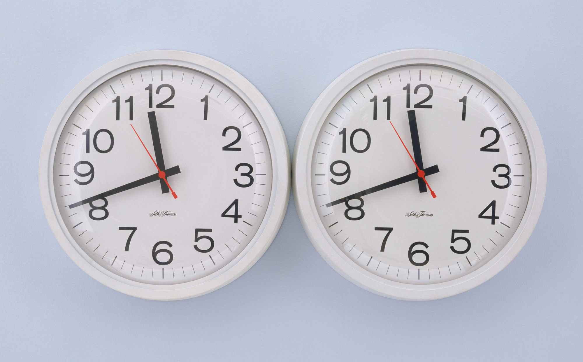 A photograph of two circular white clocks, touching side-by-side against a pale blue wall. The left clock’s hands point to 11:41:54, the other to 11:41:55